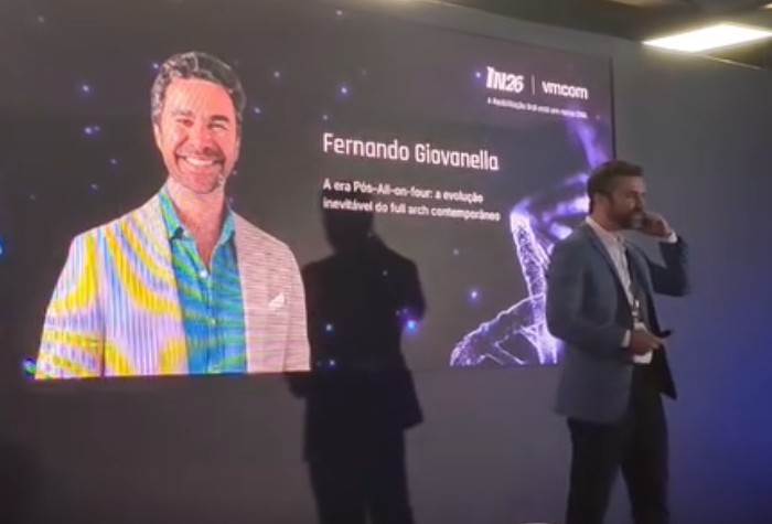 Dr. Fernando Giovanella no palco do IN26, com o seu retrato e o título da conferência no telão